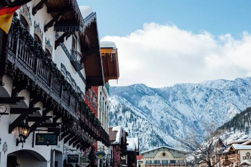 Charming alpine village with snow-covered mountains and festive decorations on buildings under a clear blue sky.