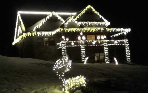 A house decorated with bright white lights and a glowing reindeer in the foreground, set against a dark night sky.