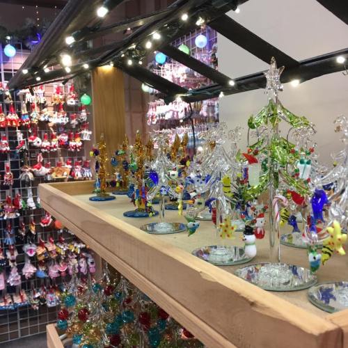 Colorful glass ornaments and decorations displayed on wooden shelves, with a festive background of hanging lights.