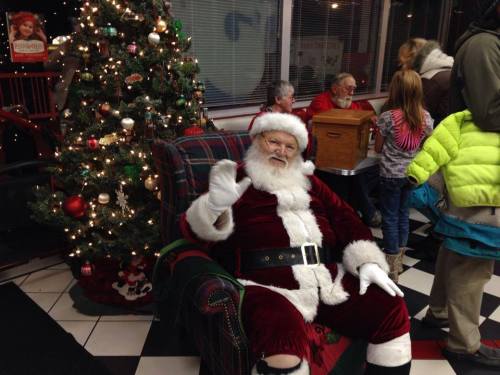 Santa Claus waves while sitting in a cozy chair near a decorated Christmas tree, surrounded by children and festive decor.