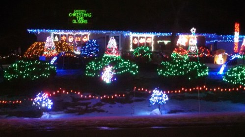 A house decorated with colorful Christmas lights, featuring trees and festive displays in a snowy yard.