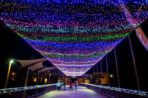 A colorful display of lights drapes over a bridge, with people walking underneath at night.