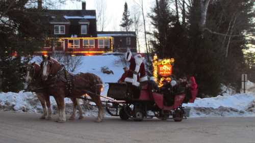 A festive sleigh with Santa and passengers, pulled by two horses, in a snowy setting near a decorated inn.