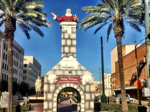 A large Santa figure atop a chimney structure, surrounded by palm trees in a festive urban setting.