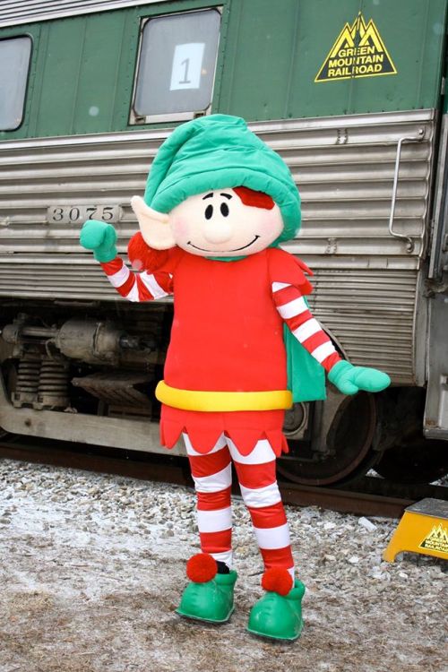 A cheerful elf mascot in a red and green outfit stands next to a train, waving in a snowy setting.