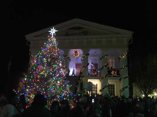 A brightly lit Christmas tree in front of a decorated building, with a crowd gathered below at night.