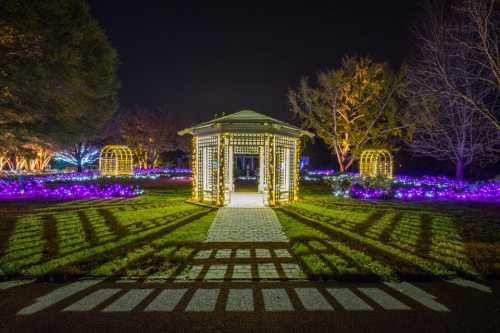 A beautifully lit gazebo surrounded by colorful lights and flowers in a nighttime garden setting.