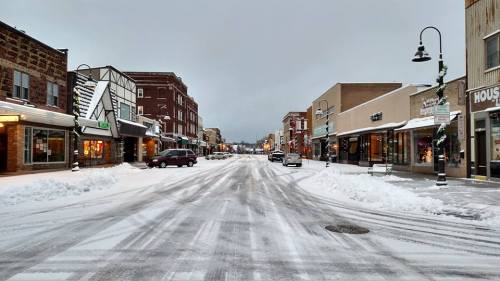 Snow-covered street lined with shops, under a gray sky, creating a quiet winter scene.