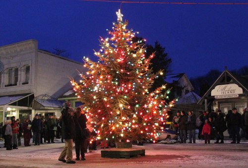 A brightly lit Christmas tree stands in a snowy town square, surrounded by people enjoying the festive atmosphere at dusk.