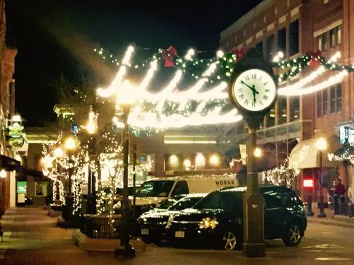 A festive street at night, adorned with holiday lights and decorations, featuring a clock and parked cars.