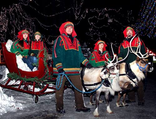 A group in festive attire stands with reindeer and a sleigh, surrounded by holiday lights and snow.