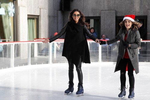 Two women ice skating at an outdoor rink, one wearing a Santa hat, both smiling and holding hands.
