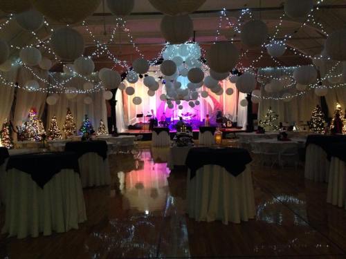 A beautifully decorated event space with string lights, paper lanterns, and Christmas trees, set for a celebration.