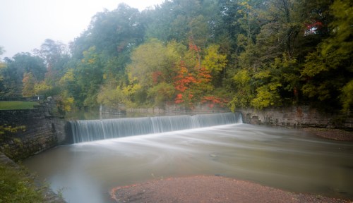 A serene waterfall cascades over a stone dam, surrounded by vibrant autumn foliage in soft, misty light.