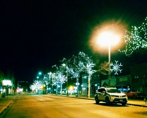 A quiet street at night adorned with festive white lights on trees and buildings, illuminated by street lamps.