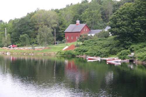 A serene lake reflects a red barn and surrounding greenery, with boats docked along the shore.