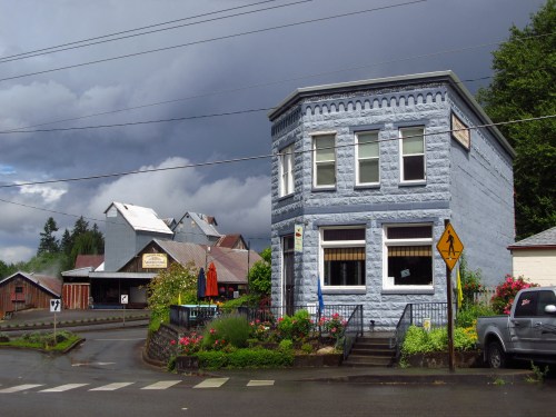 A blue historic building with flowers in front, set against a cloudy sky and nearby rustic structures.