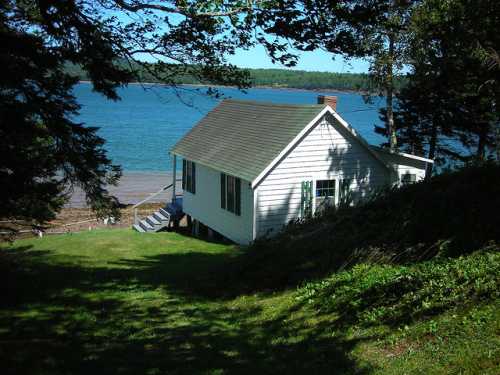 A small white cottage sits on a grassy slope by a blue lake, surrounded by trees and a clear sky.