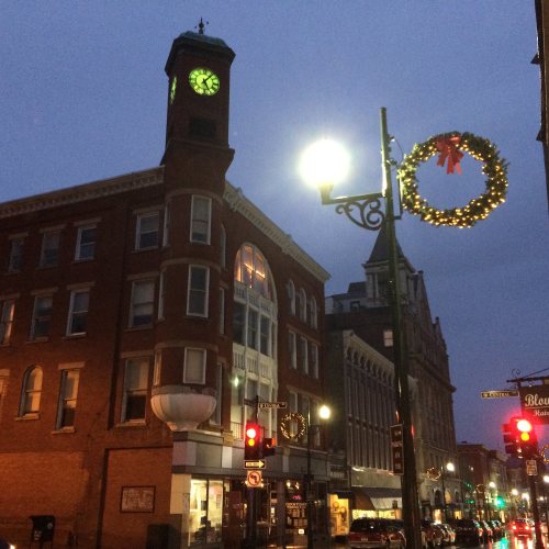 Historic buildings adorned with holiday decorations, a clock tower, and streetlights glowing at dusk.
