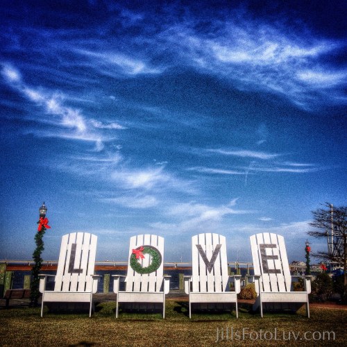 Four white chairs arranged to spell "LOVE," decorated with a wreath, against a blue sky and festive atmosphere.