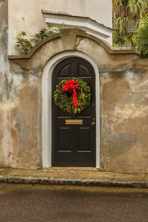 A black door with an arched top, adorned with a festive wreath and red bow, set against a weathered wall.