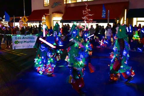 A festive parade scene with participants dressed as Christmas trees, adorned with lights and decorations, celebrating the holiday.