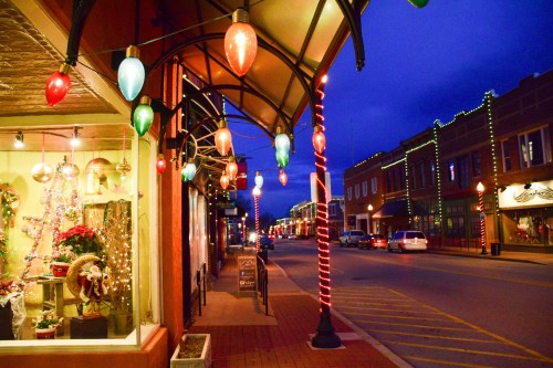 A festive street scene at dusk, adorned with colorful holiday lights and decorations on buildings and storefronts.