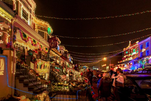 A festive street scene with houses adorned in colorful holiday lights and people enjoying the decorations at night.