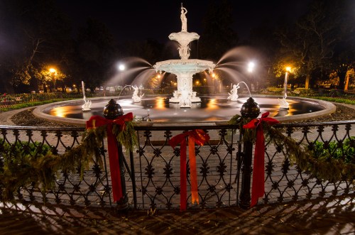 A beautifully lit fountain at night, surrounded by festive decorations and greenery, with flowing water and statues.