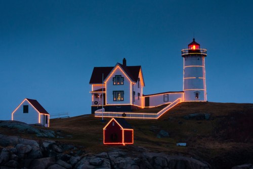 A lighthouse and house decorated with glowing lights at dusk, surrounded by rocky terrain.