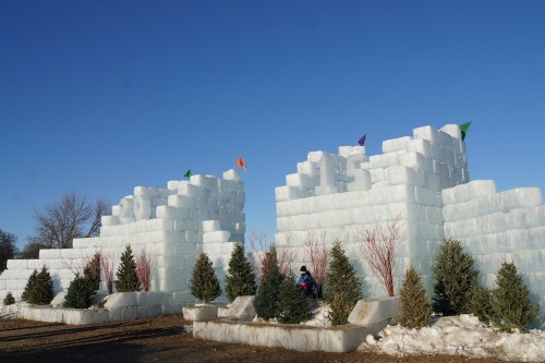 A large ice castle with colorful flags, surrounded by evergreen trees, under a clear blue sky.