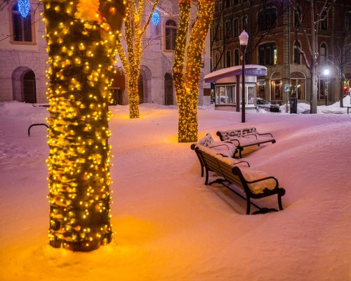 A snowy scene with trees wrapped in warm lights and benches covered in snow, creating a cozy winter atmosphere.