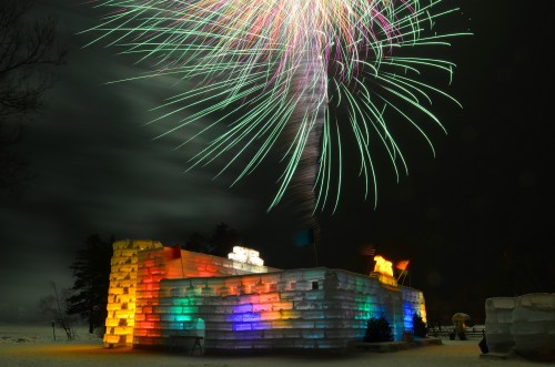 A colorful ice structure illuminated at night, with vibrant fireworks bursting above it in a dark sky.