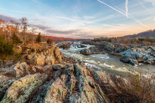 A scenic view of a river flowing over rocky terrain, surrounded by trees and a colorful sky at sunset.