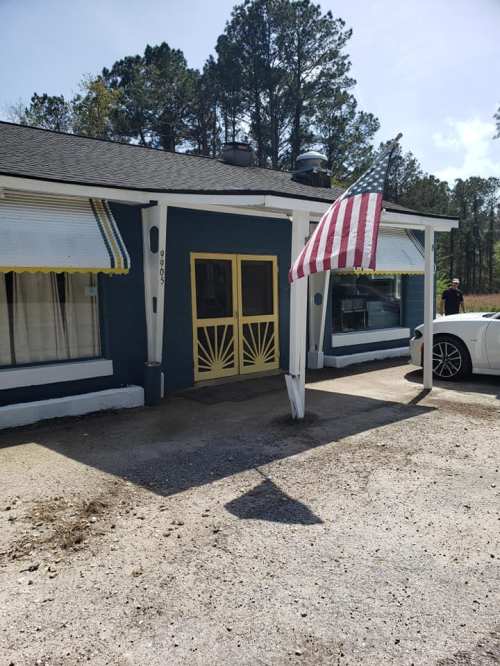 A blue house with yellow trim and an American flag in front, surrounded by trees and a gravel driveway.