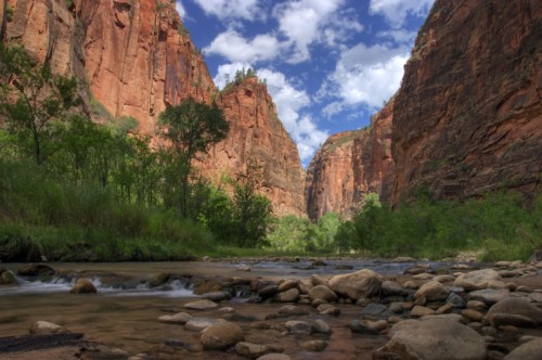 A scenic view of a canyon with towering red rock walls, lush greenery, and a flowing river in the foreground.
