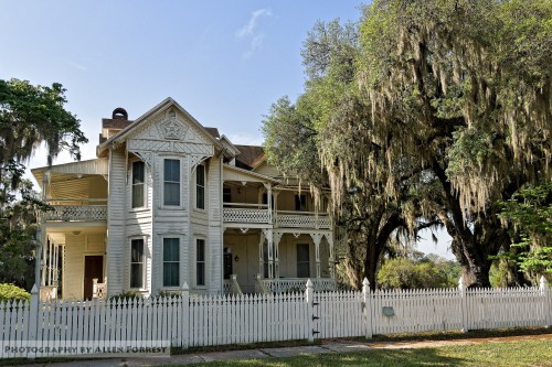 A historic two-story house with a wraparound porch, surrounded by trees and a white picket fence.