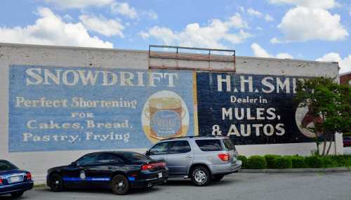 Faded mural on a brick wall advertising Snowdrift shortening and H.H. Smith, dealer in mules and autos, with parked cars below.