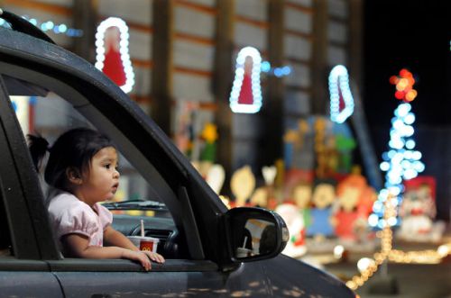 A young child leans out of a car window, gazing at colorful holiday lights in the background.