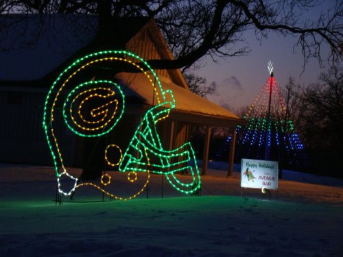 A glowing green and yellow football helmet decoration next to a colorful Christmas tree and a holiday sign in the snow.