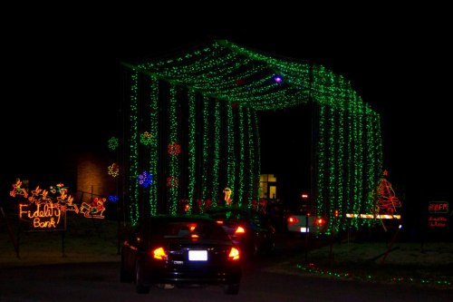A festive display of green holiday lights creating an archway, with colorful decorations and cars passing through at night.