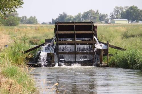 A wooden water wheel structure in a stream, with water cascading over it surrounded by lush greenery.
