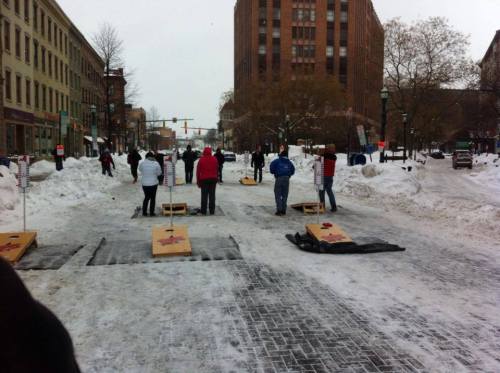 People playing cornhole on a snowy street, surrounded by snowbanks and buildings, with a cloudy sky above.