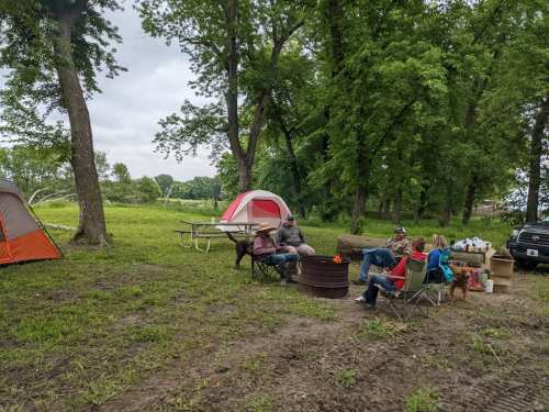 A group of people sitting around a campfire near tents and a picnic table in a wooded area.