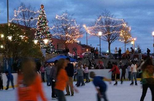 A festive outdoor ice skating rink with a decorated Christmas tree and twinkling lights, filled with skaters and spectators.
