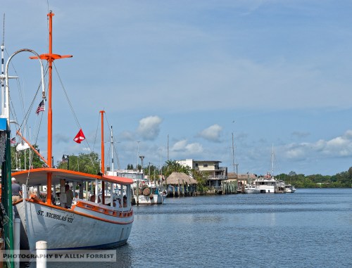 A scenic view of a harbor with a boat named "St. Nicholas IV" and buildings along the waterfront under a blue sky.