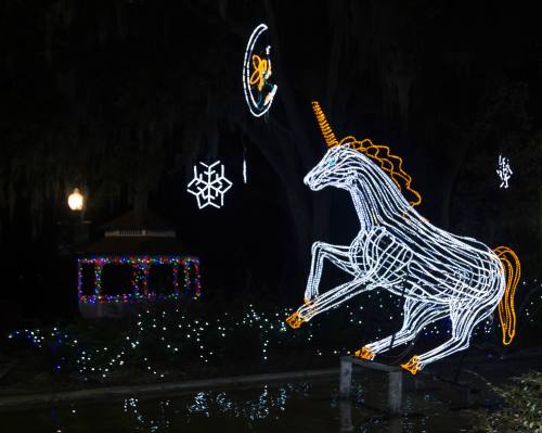 A glowing white unicorn made of lights leaps in a dark park, surrounded by festive decorations and twinkling lights.