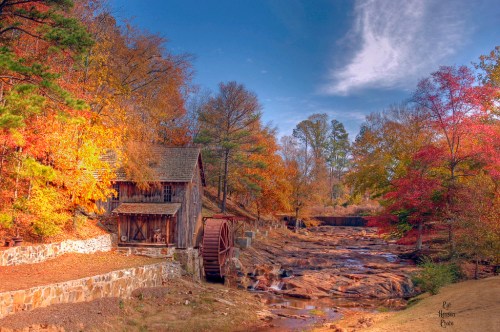 A scenic autumn landscape featuring a rustic mill beside a rocky stream, surrounded by colorful fall foliage.