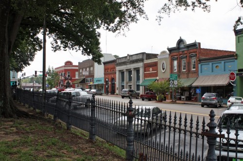 A quaint town street lined with shops, cars, and a decorative iron fence under a cloudy sky.