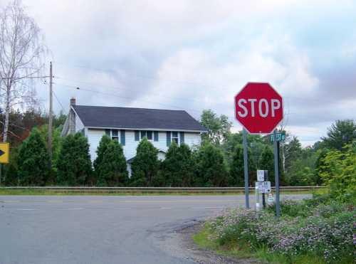 A stop sign at an intersection with a house and trees in the background under a cloudy sky.